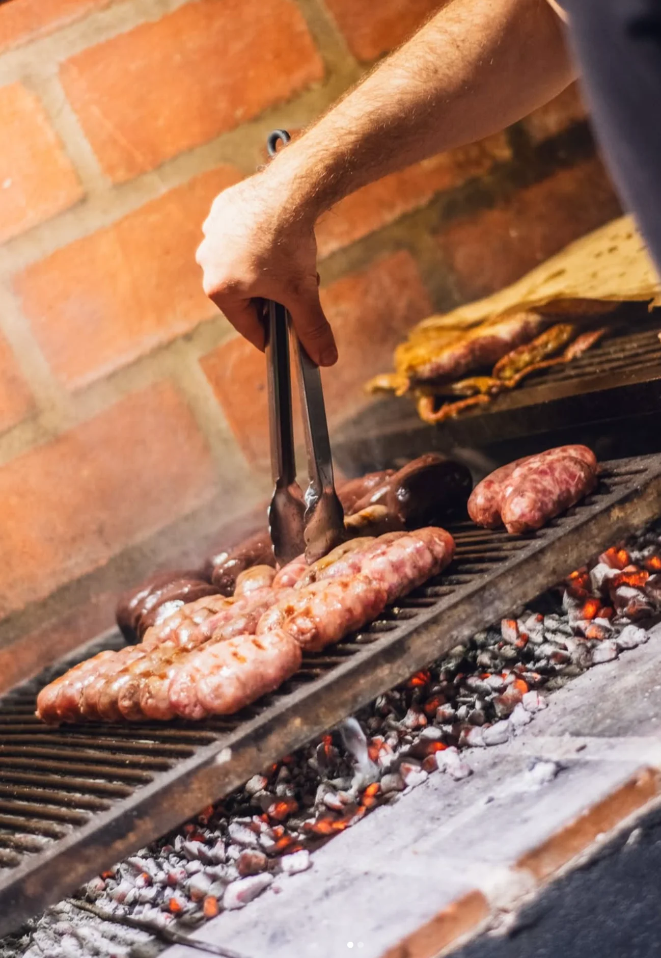 Parrillero dando vuelta chorizos y morcillas con la pinza, sobre brasas encendidas